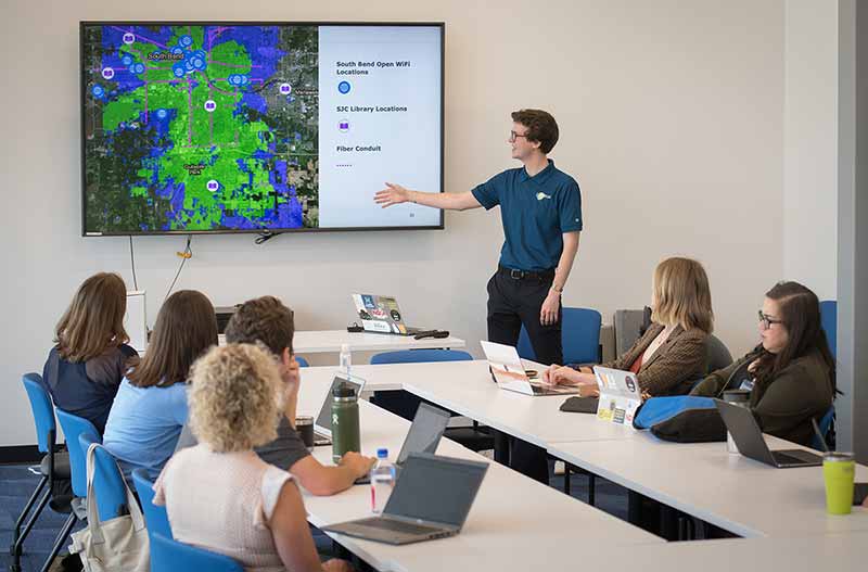 Patrick McGuire, a man wearing glasses and a blue polo shirt, stands in front of a small ground sitting around a table. He points at a large screen displaying a map of open wifi in a region.
