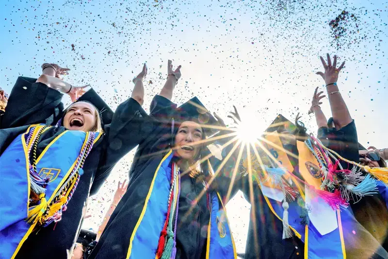 A group of student throwing confetti in the air. The sun is shining brightly in the background.