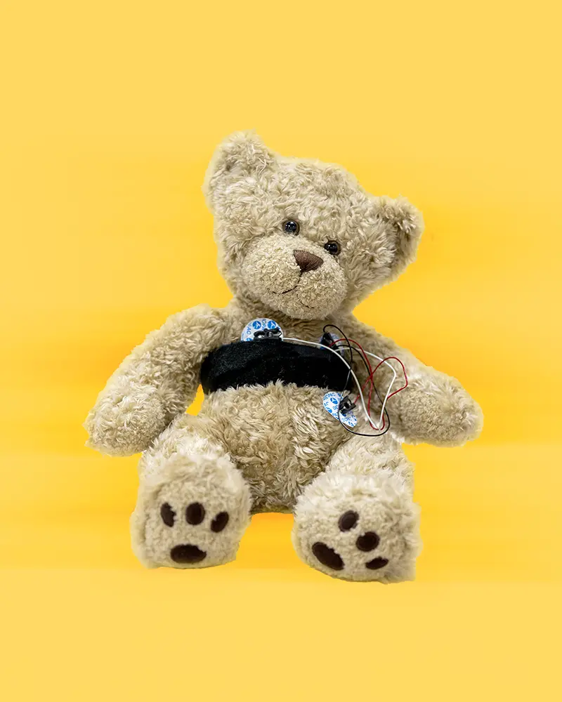 A teddy bear with with electrode sensors attached to it on a blurry, yellow background.