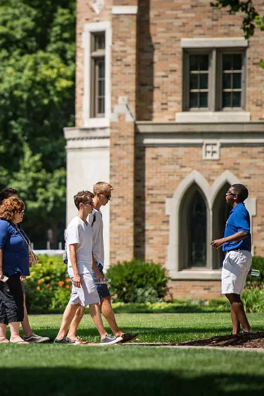 A diverse group of college students walks on a sunny green lawn with a brick building in the background at the University of Notre Dame.