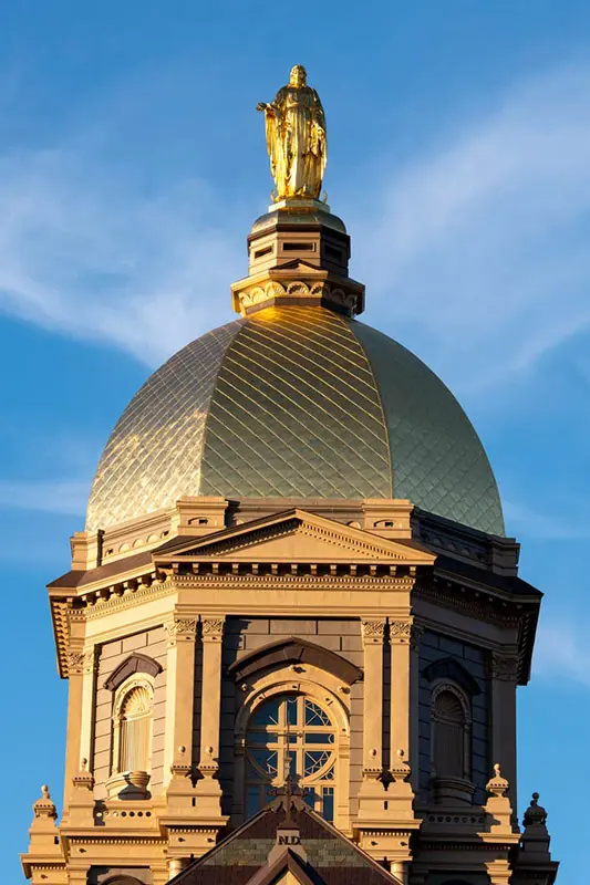The golden dome and a statue of Mary at the Main Building of the University of Notre Dame are visible against a clear blue sky.