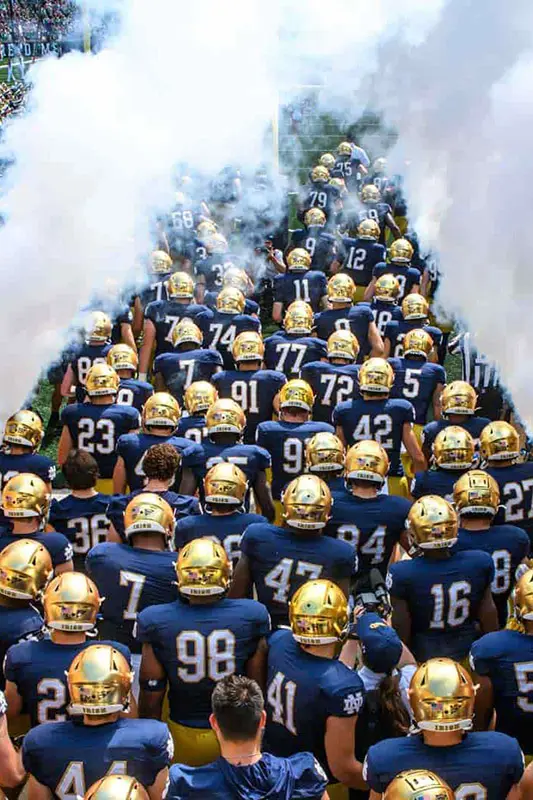 The Notre Dame Fighting Irish football team, in blue uniforms and gold helmets, runs onto the field through a cloud of smoke from an overhead perspective.