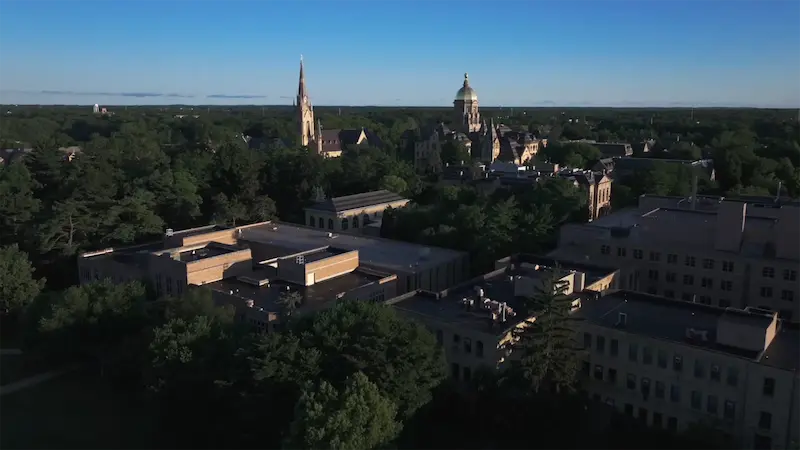 An aerial view of the University of Notre Dame campus shows the Golden Dome and Sacred Heart Basilica rising above a tree-lined landscape under a clear blue sky.