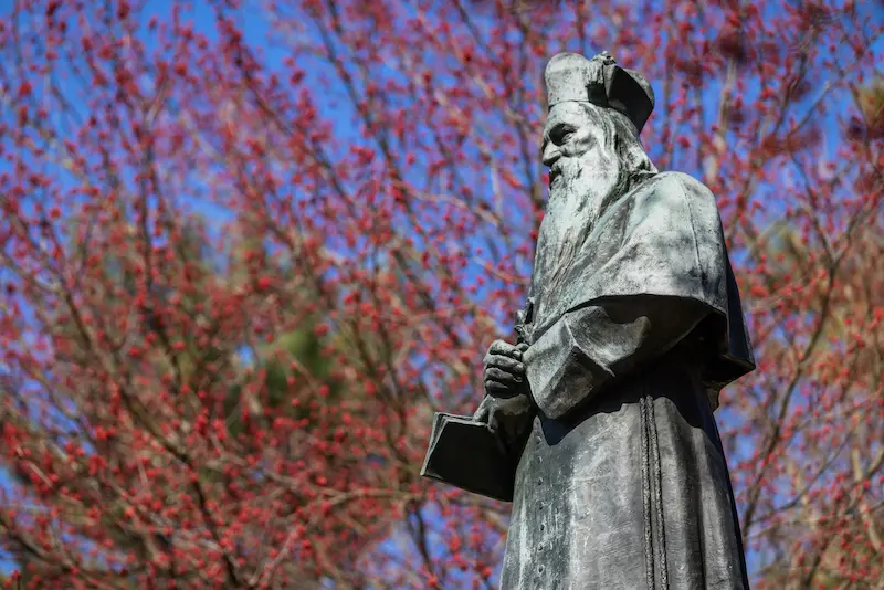 A statue of Rev. Edward Sorin, C.S.C., founder of the University of Notre Dame, stands against a bright blue sky and a tree with red foliage.