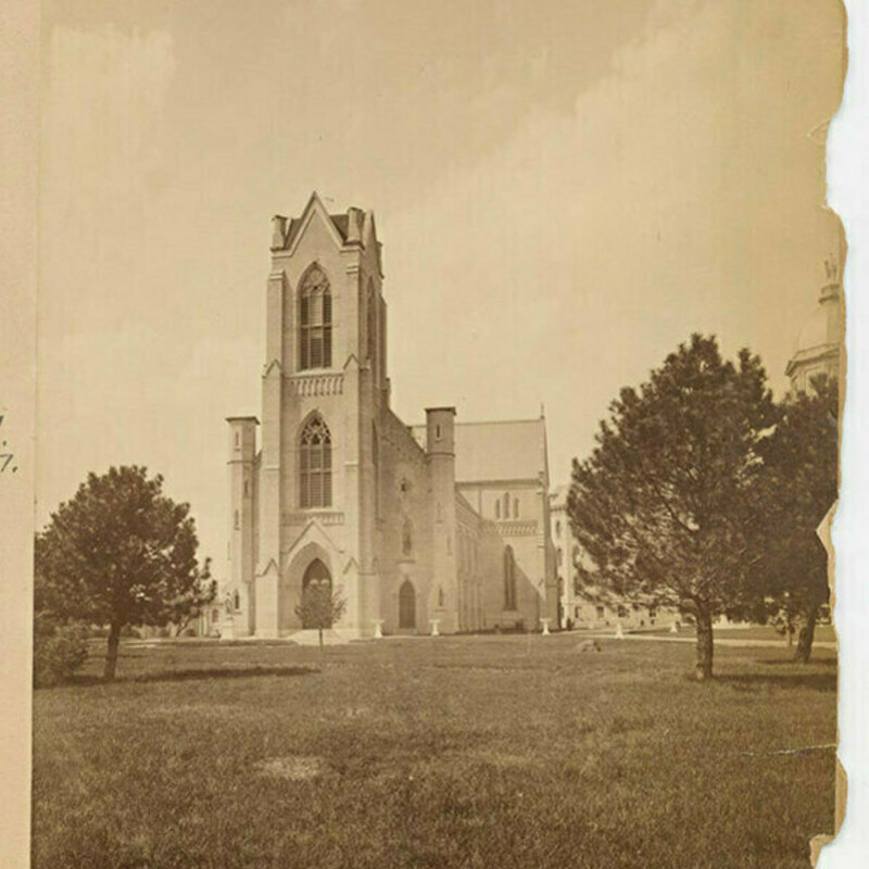 Sepia-toned photo of the Church of the Sacred Heart at the University of Notre Dame in 1885. The church's facade, with its pointed arches and tall central tower, is seen across a grassy lawn with young trees.