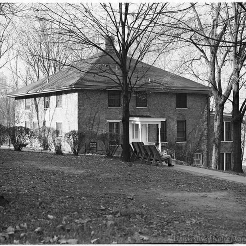 Black and white photo of Sorin Hall at the University of Notre Dame. A person sits in a wooden Adirondack chair in front of the brick building, facing away from the camera. Bare trees and a leaf-covered lawn surround the building.