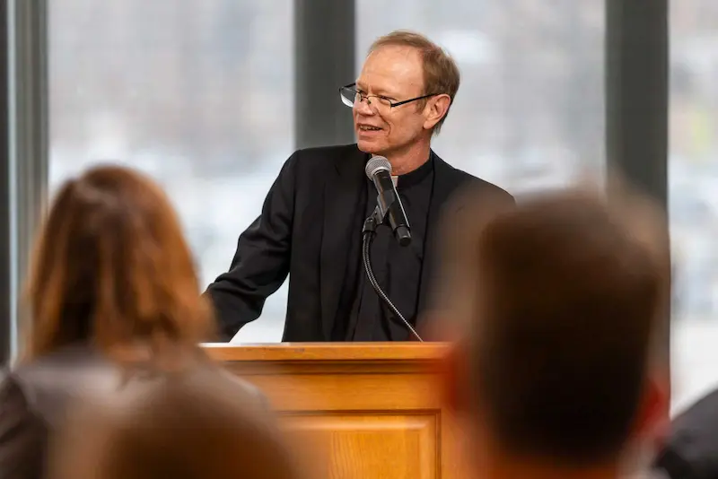 Father Dowd speaks at a wooden podium with a microphone, wearing a black suit and clerical collar.