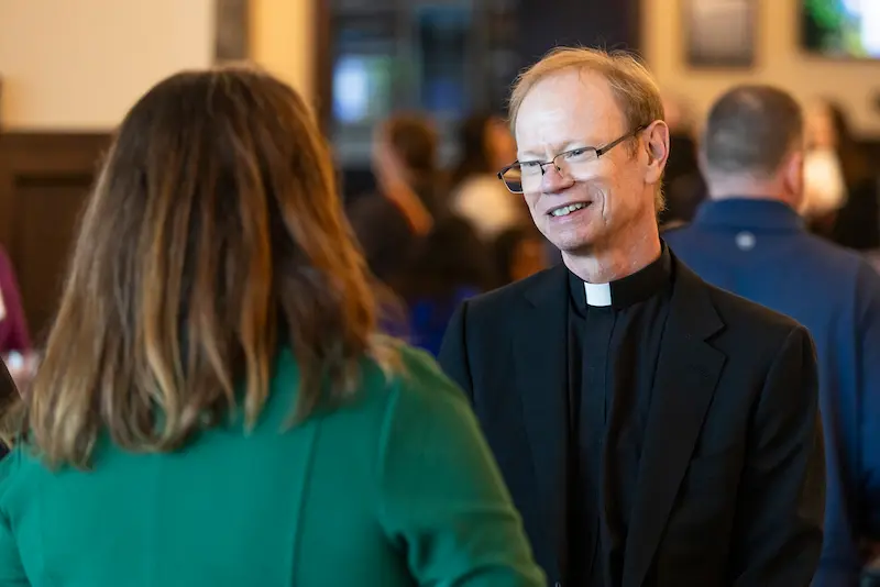 President Rev. Robert A. Dowd, C.S.C., in a black suit and clerical collar, smiles while speaking with a woman in a green jacket.