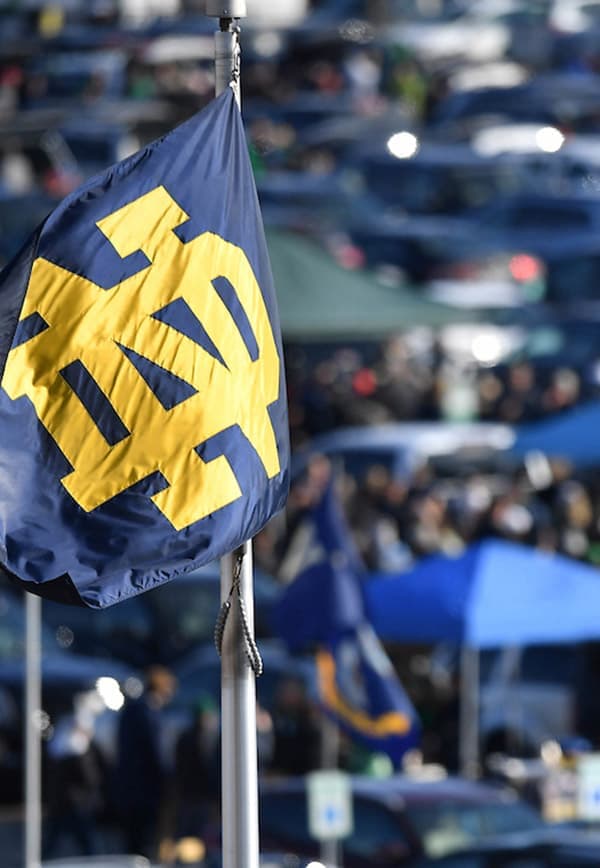 A blue flag with a yellow interlocking “ND” logo flies above a crowded parking lot with tents and vehicles in the background.