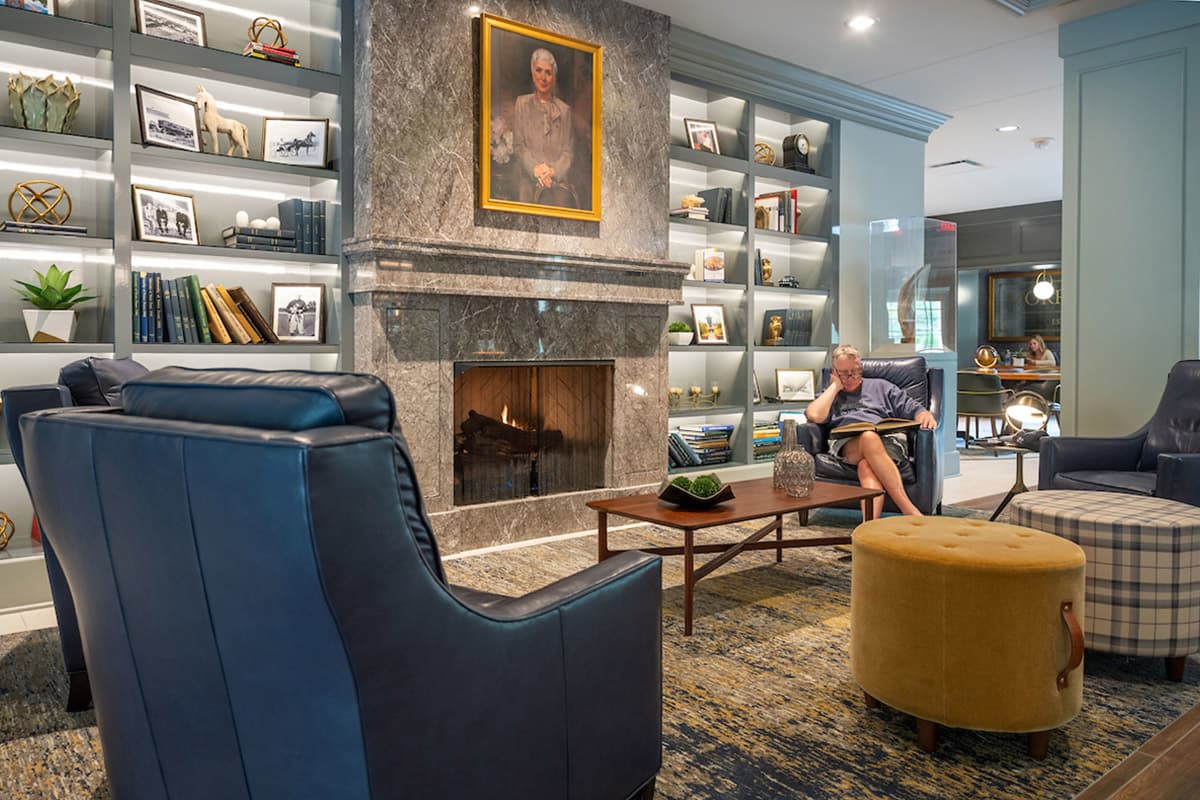 Cozy lounge with a marble fireplace, blue leather chairs, and bookshelves filled with decor. A person reads a book, creating a peaceful atmosphere.