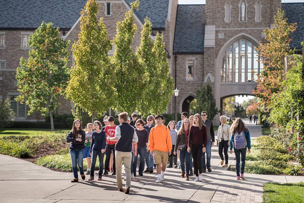 A group of students tours campus on a sunny day. They walk along a path with green trees and the law school in the background.