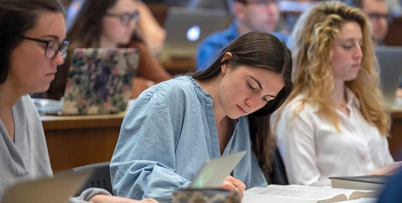A female student with long brown hair wears a light blue denim shirt and reads a book in a classroom, with other students visible in the background.