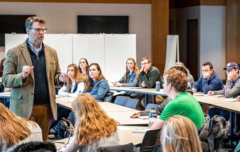 A male instructor in a tweed jacket and glasses stands at the front of a classroom, gesturing as he speaks to seated students.