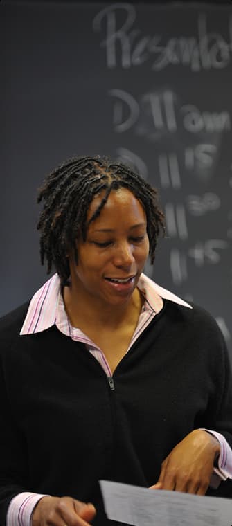 A Black woman with dreadlocks wears a black zippered sweater over a striped shirt, holding papers and speaking in front of a chalkboard.