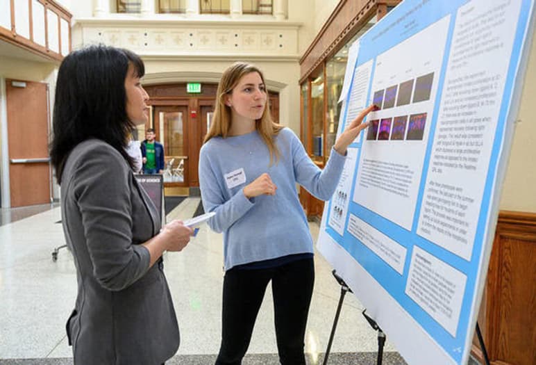 A female student with long blonde hair in a light blue sweater points to a large poster board display for another woman in a gray suit jacket.