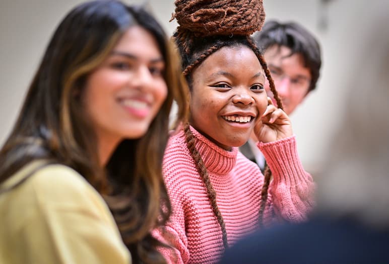 Two smiling students are in a classroom. One student has a light brown complexion and long dark hair, and the other student has dark skin with long dreadlocks and is wearing a pink sweater.