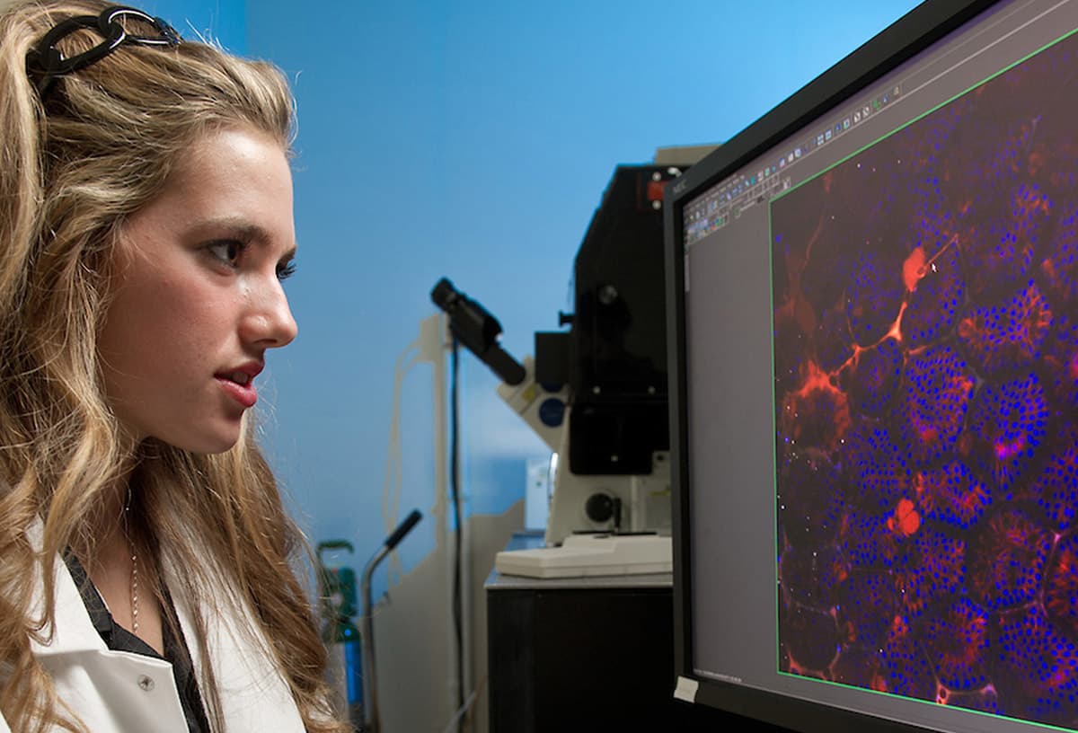 A young woman in a white lab coat looks intently at a computer screen displaying a microscopic image in shades of red and blue, with a microscope in the background.
