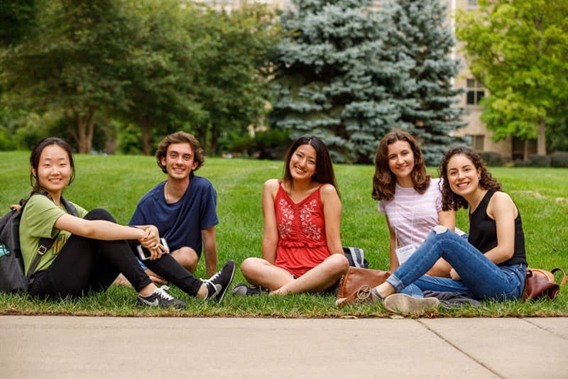 Five students, one male and four females, are seated on a grassy lawn with a building and pine trees in the background, all smiling at the camera.
