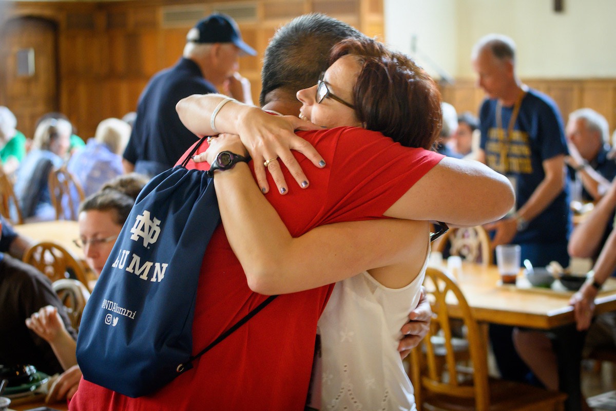 A woman in glasses and a white tank top hugs a man wearing a red shirt and a blue Notre Dame alumni drawstring bag on his back in a crowded room.