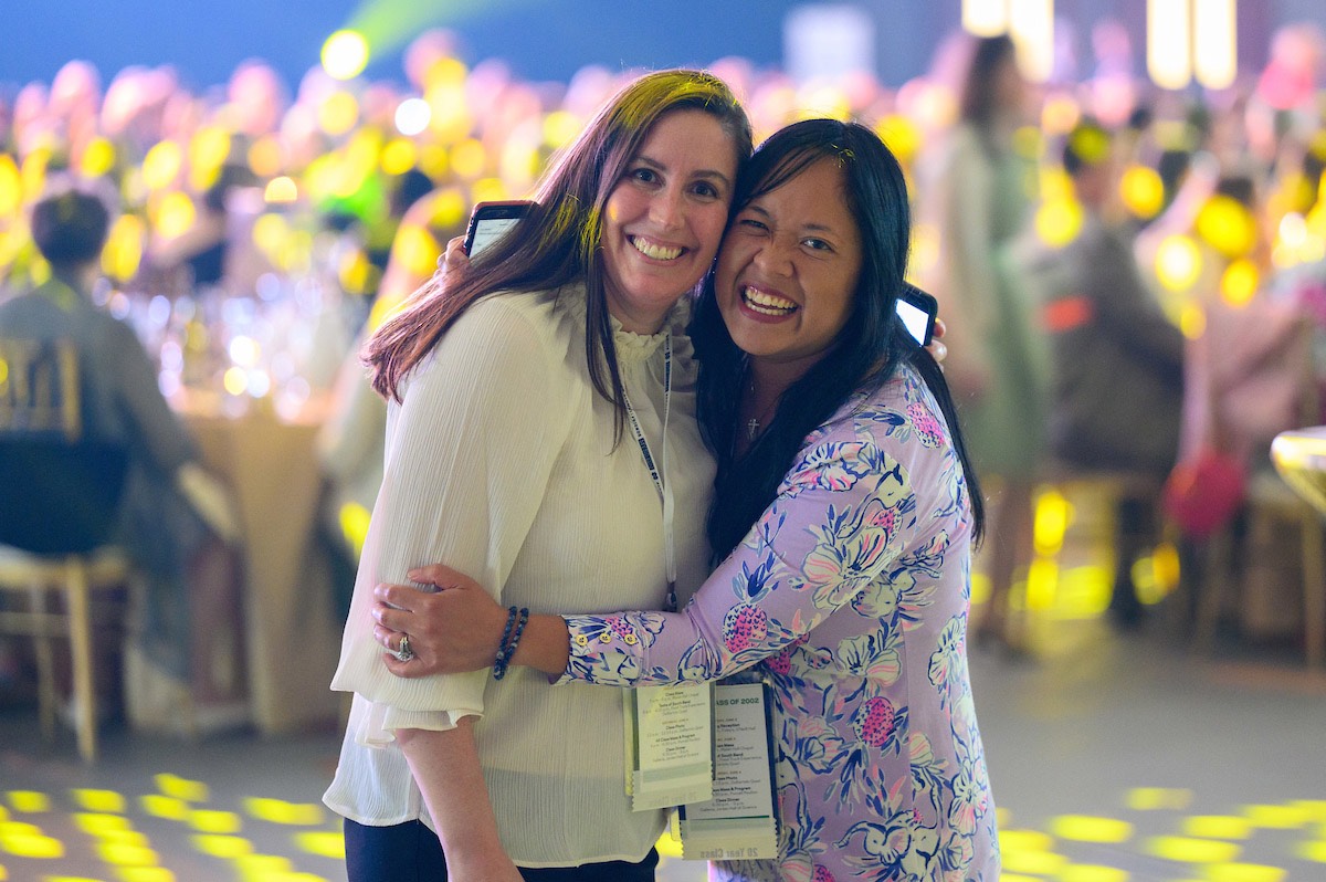Two smiling women embrace, one with long brown hair in a white shirt and the other with dark hair in a floral purple top, at an indoor event.