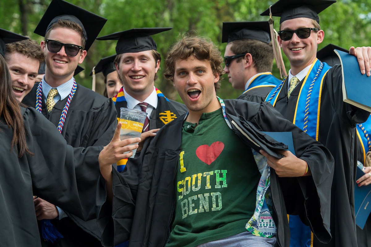A graduating student in a cap and gown reveals a green “I Love South Bend” t-shirt, surrounded by other smiling graduates.