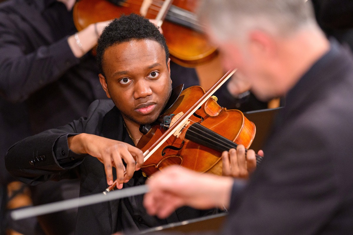 A young Black man with a serious expression plays the violin in an orchestra, looking towards the conductor's blurred back.