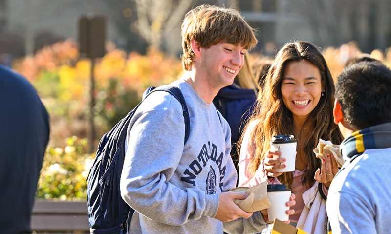 A diverse group of smiling college students, one wearing a Notre Dame sweatshirt, hold coffee cups and snacks outdoors on a sunny day.