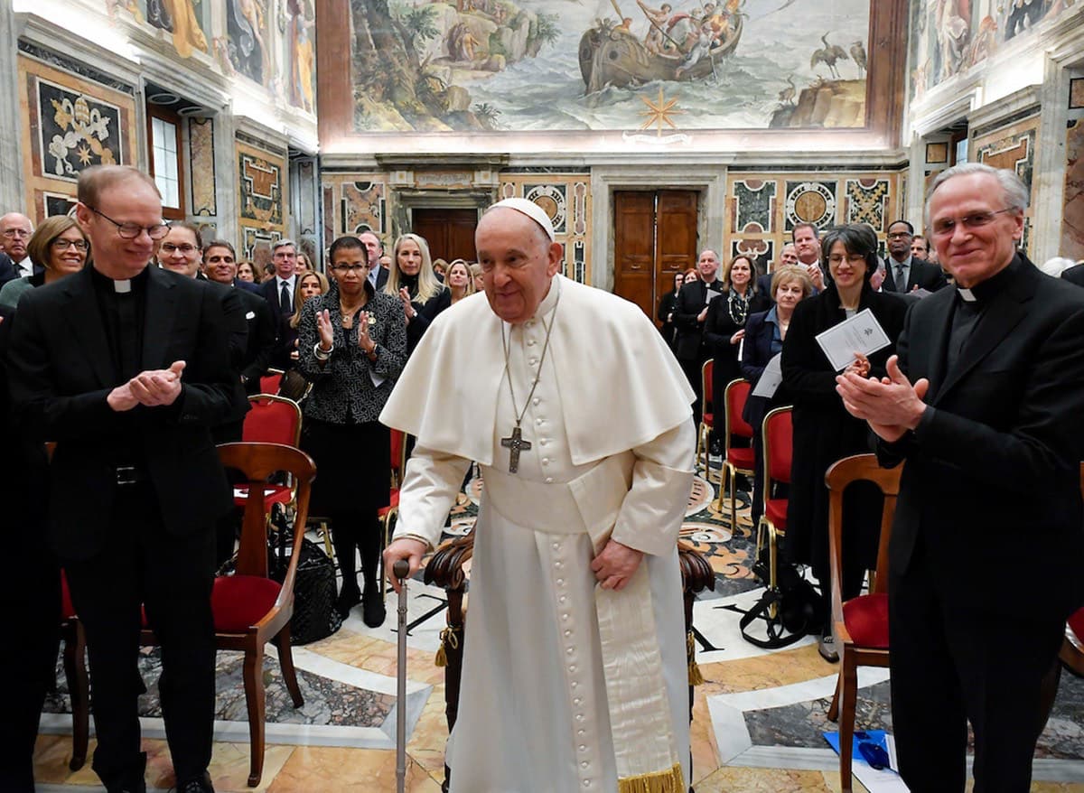 Pope Francis, dressed in white, walks with a cane in a large room with painted walls, flanked by Fathers Dowd and Jenkins and an applauding crowd.