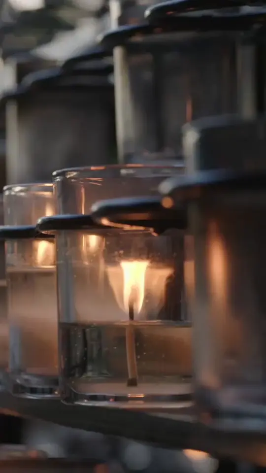 A close-up shows several votive candles in glass holders on a rack, with one flame burning brightly.