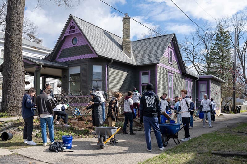 A group of college students works in the yard of a dark gray house with purple trim, using tools and wheelbarrows for a service project.