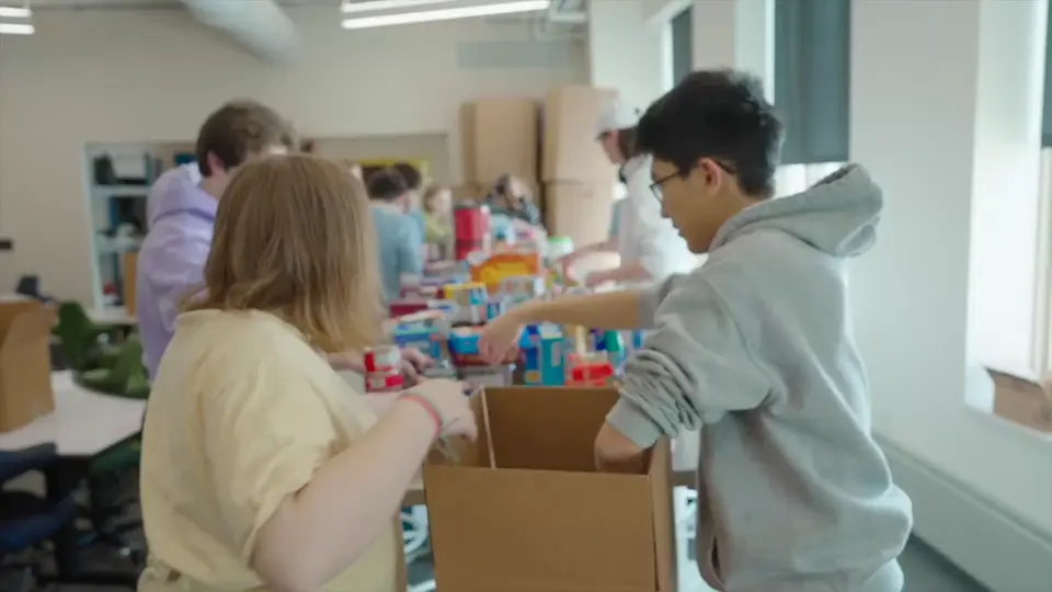Two students pack canned goods and other items into cardboard boxes at a long table in a bright room, with other students in the background.