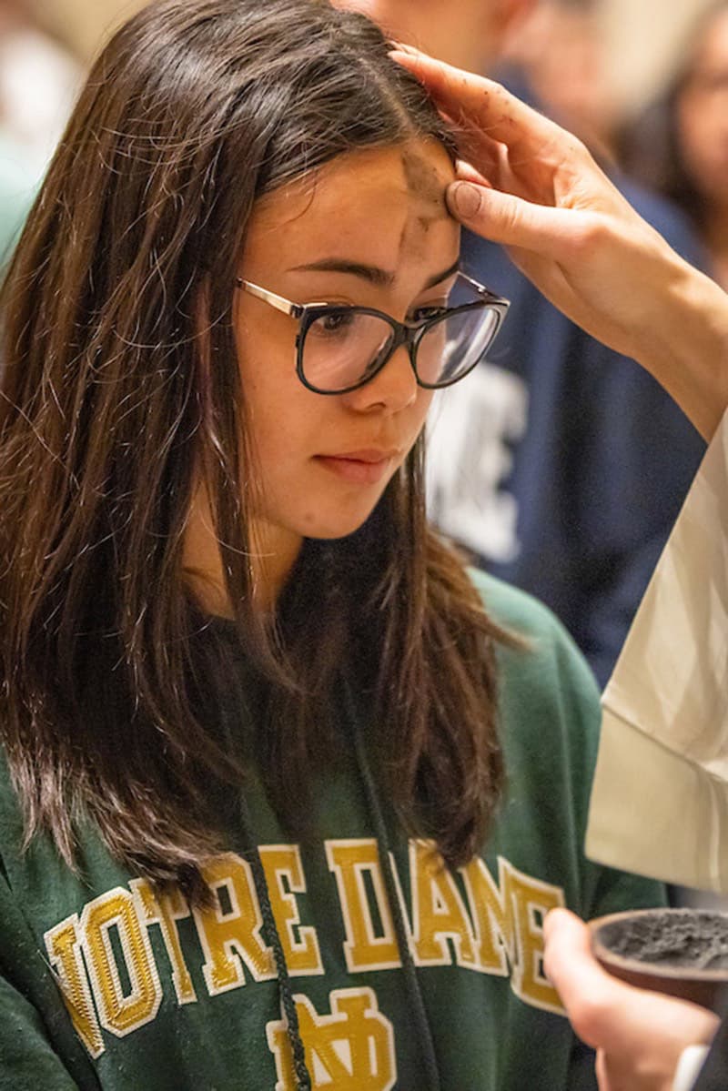 A person with long brown hair and glasses receives ashes on their forehead during an Ash Wednesday service.