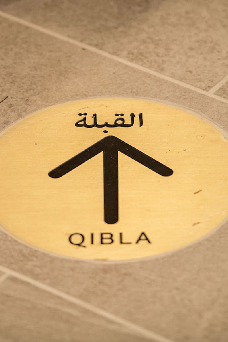 A yellow circular floor sign with an arrow and the word “QIBLA” in English and Arabic points the direction of prayer.