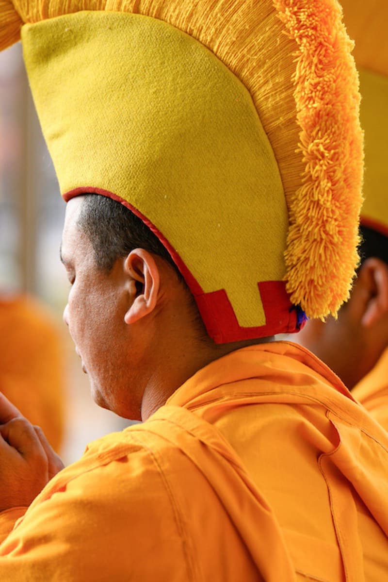A person in orange robes wears a distinctive yellow and red hat with a fringe, seen from a side and slightly back angle.