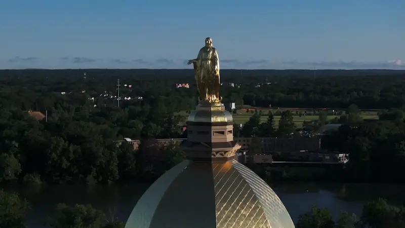 The golden dome and statue of Mary at the Main Building of the University of Notre Dame rise above green trees and a river under a clear blue sky.