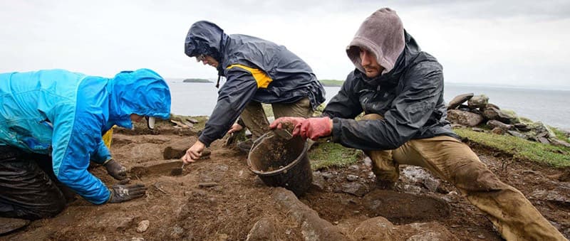 Three people in rain gear are actively digging in muddy earth, with one holding a bucket, near a body of water under an overcast sky.