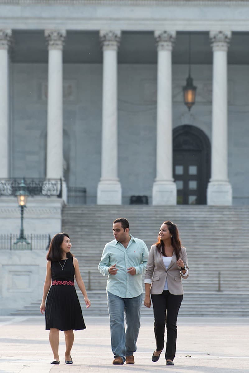 Three people, two women and one man, walk and talk in front of a large neoclassical building with columns and steps.