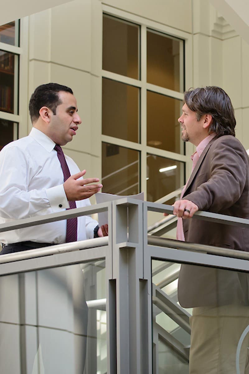 Two men, one in a white shirt and tie and the other in a brown jacket, stand on a balcony and engage in conversation.