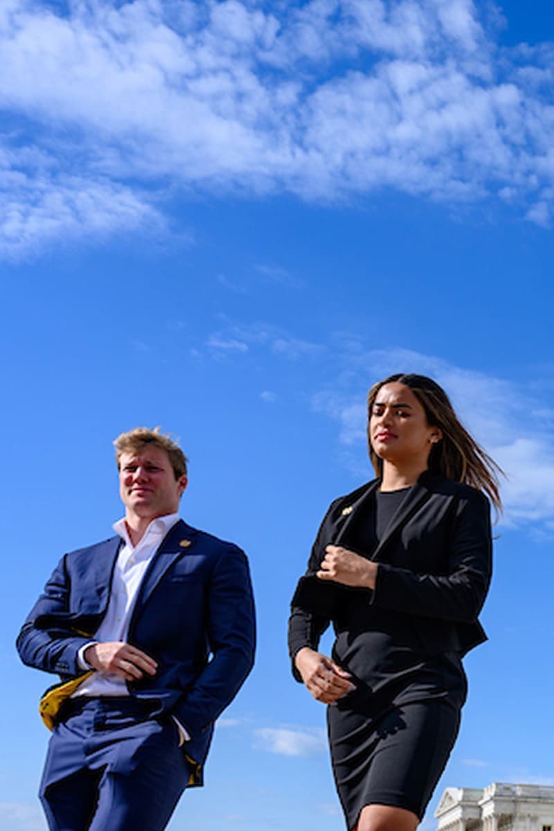 A man in a blue suit and a woman in a black dress stand outdoors, looking contemplative against a bright blue sky with wispy clouds.