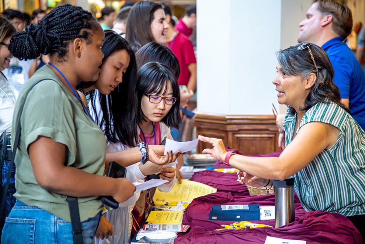 A woman with graying hair at a table explains something to three young women, one wearing glasses, at a bustling indoor event.