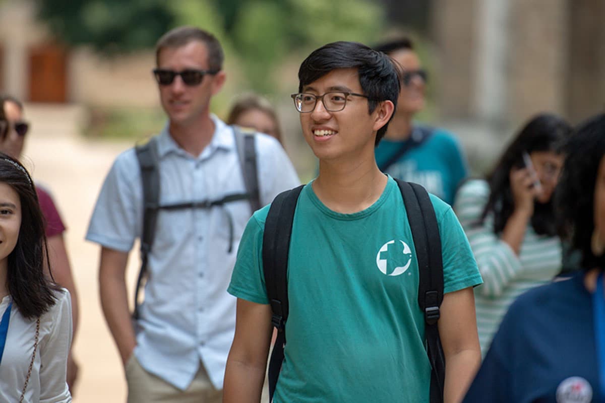 A young man in glasses and a teal t-shirt with a white circular logo smiles while walking outdoors with other students.