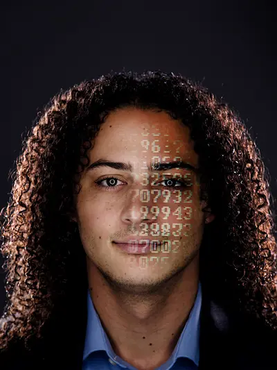 A person with curly brown hair looks directly ahead, with digits projected onto their face, specifically over one eye and across their cheek.