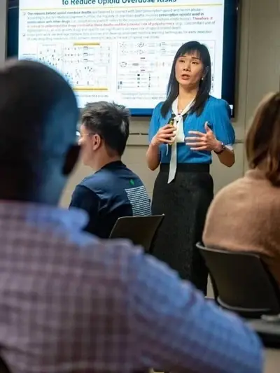 A professor stands in front of a screen displaying a presentation on opioid risks, speaking to a seated class of students.