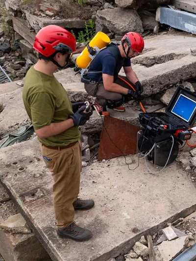 Two people in red helmets examine equipment amidst rubble.