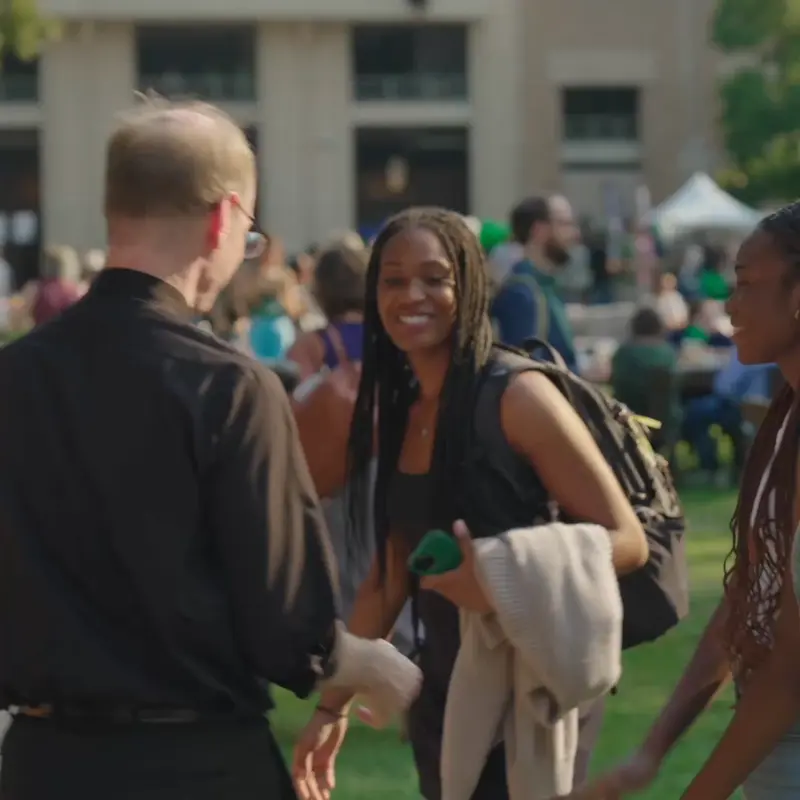 A priest in a black clerical shirt smiles and talks with two young women outdoors on a sunny day, with a crowd of people and buildings in the background.