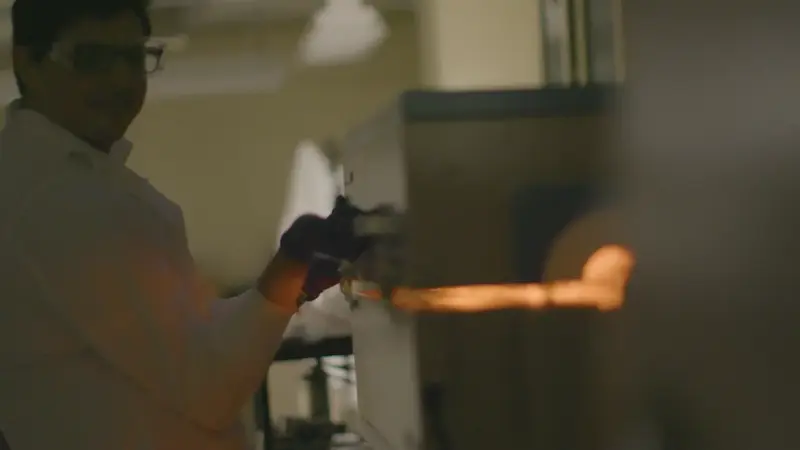 A blurred scientist in safety glasses and a lab coat is seen with gloved hands holding an object glowing orange inside a furnace.