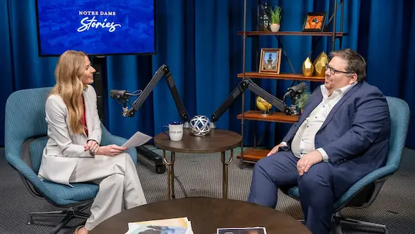 A woman interviews a man in a studio, both seated in teal chairs.
