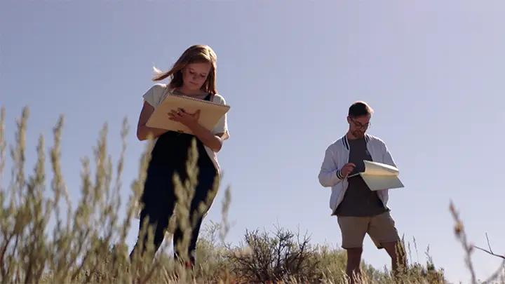 Two researchers, a woman with long blonde hair and a man with glasses, stand in a sunny, grassy field taking notes on clipboards.
