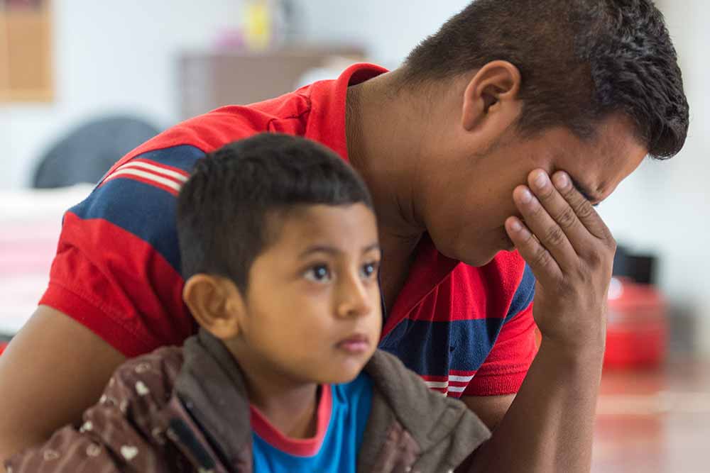 Jose holding his head in his hand as his son sits on his lap.