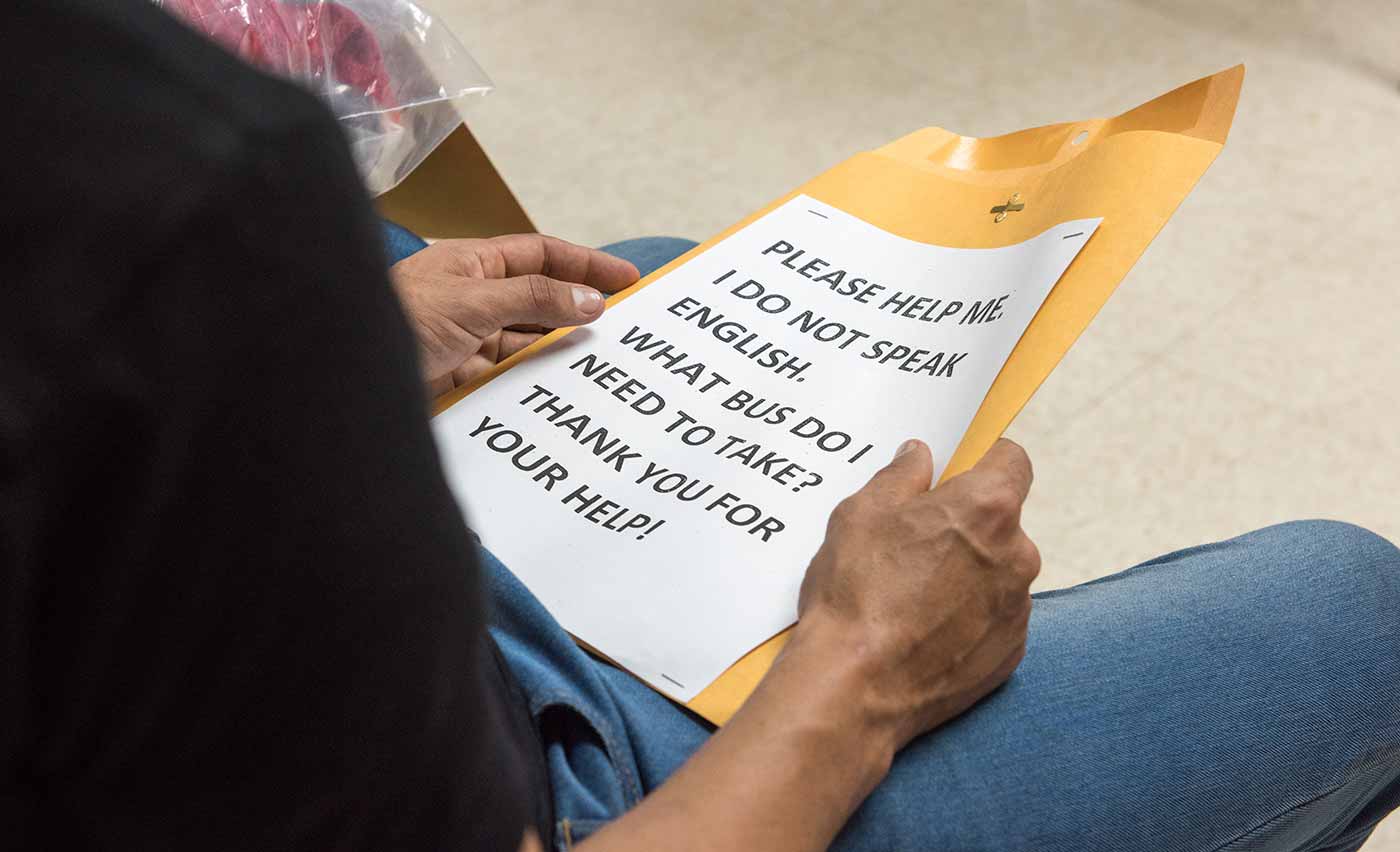 A man sitting on a chair with a sign reading “Please help me. I do not speak English. What bus do I need to take? Thank you for your help!”